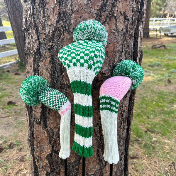 Knitted golf club covers in green, white, and pink against a tree trunk.
